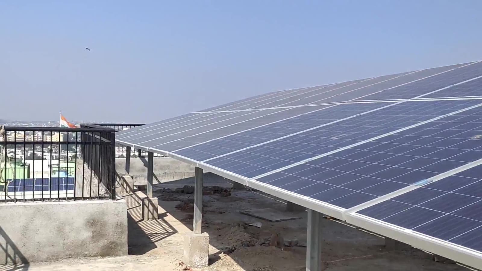 Rows of solar panels on an elevated steel structure on a rooftop, with a railing and the city skyline beyond