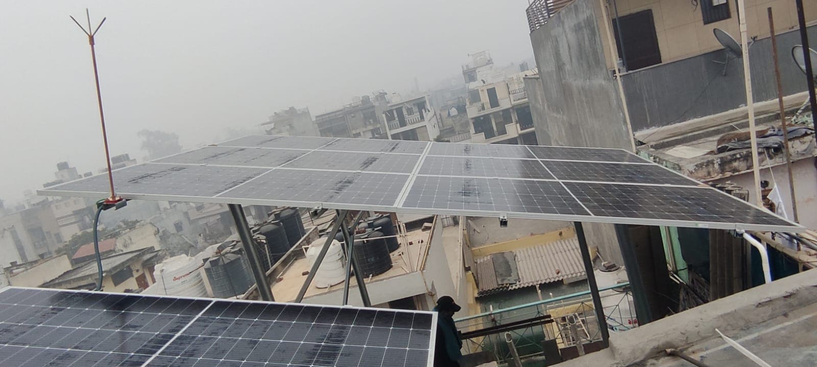 Solar panels on a tall elevated steel structure above a rooftop with a hazy city skyline behind