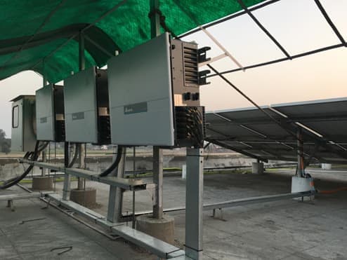 Three grey string inverters mounted side by side on a steel frame under a green shade net, with solar module rows behind them on a flat roof