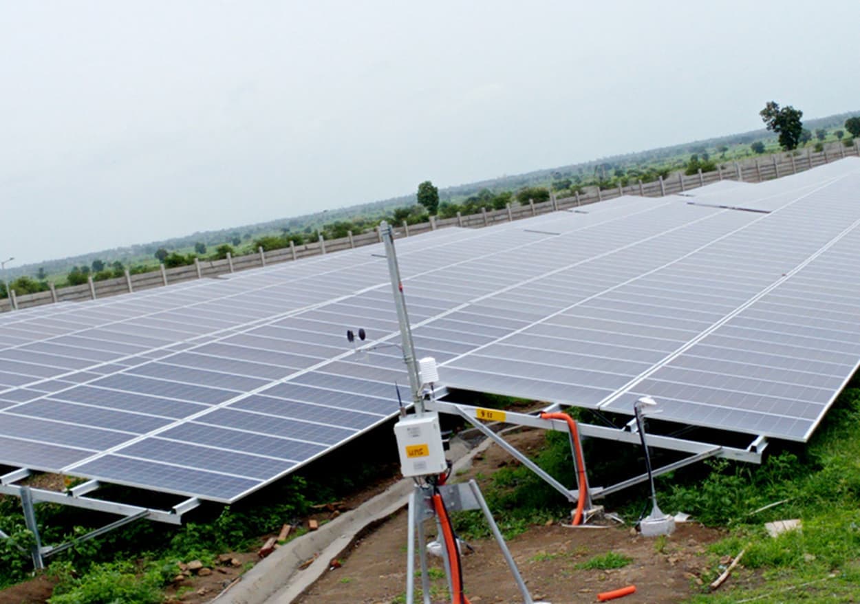 Ground-mounted solar array in open farmland with a weather monitoring mast in the foreground under an overcast sky