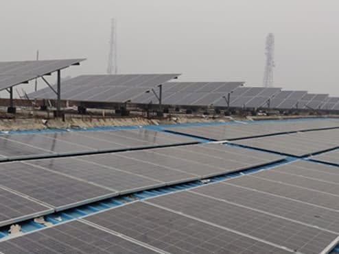 Rows of solar panels on a flat factory roof, with further panel rows and two lattice towers behind them under a hazy sky