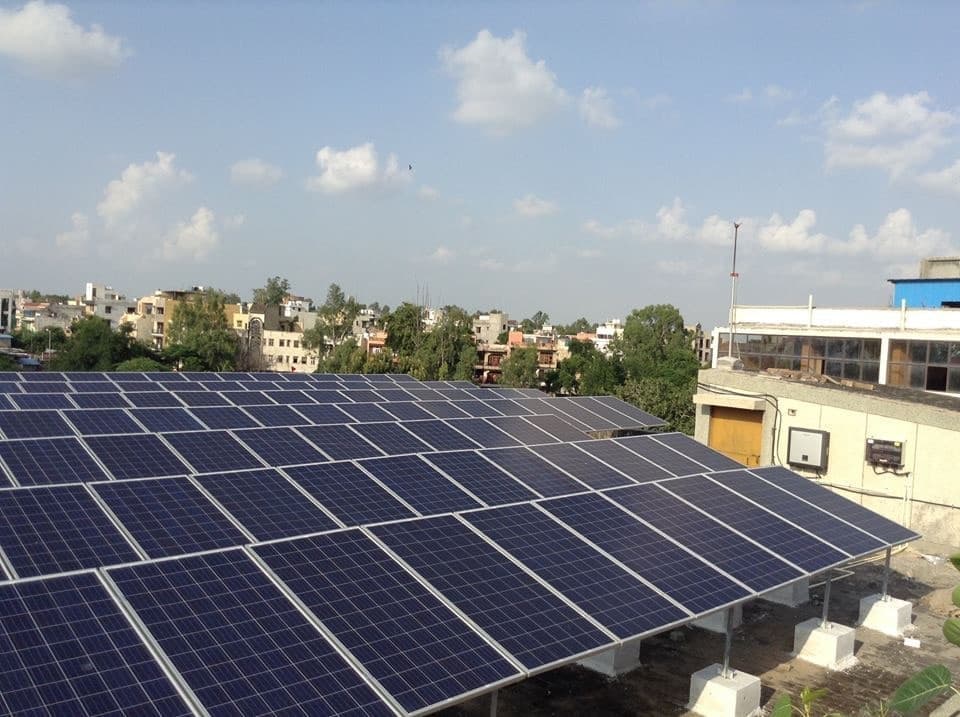 Wide rooftop array of tilted solar panels on concrete footings with trees and city buildings under a partly cloudy sky