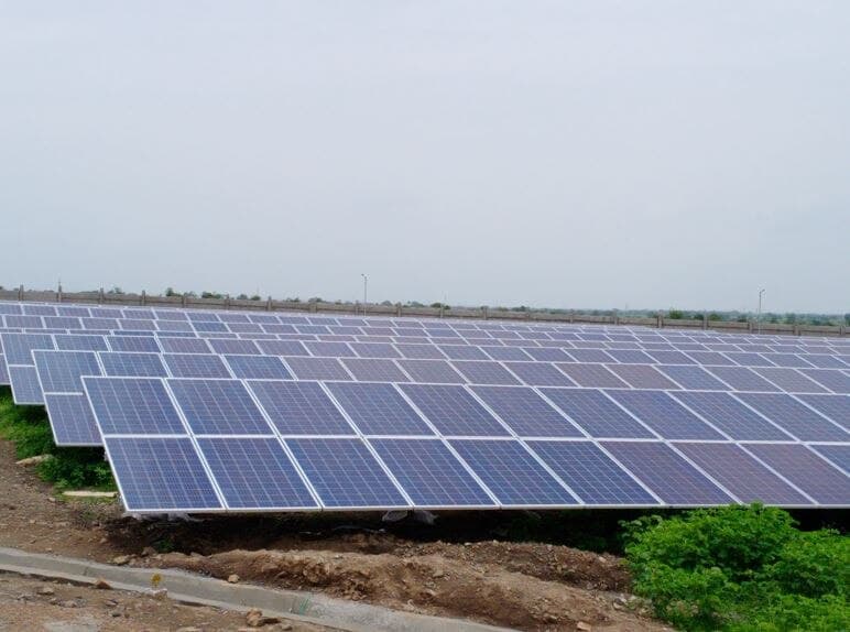 Rows of ground-mounted solar panels on a flat plot of red earth under an overcast sky