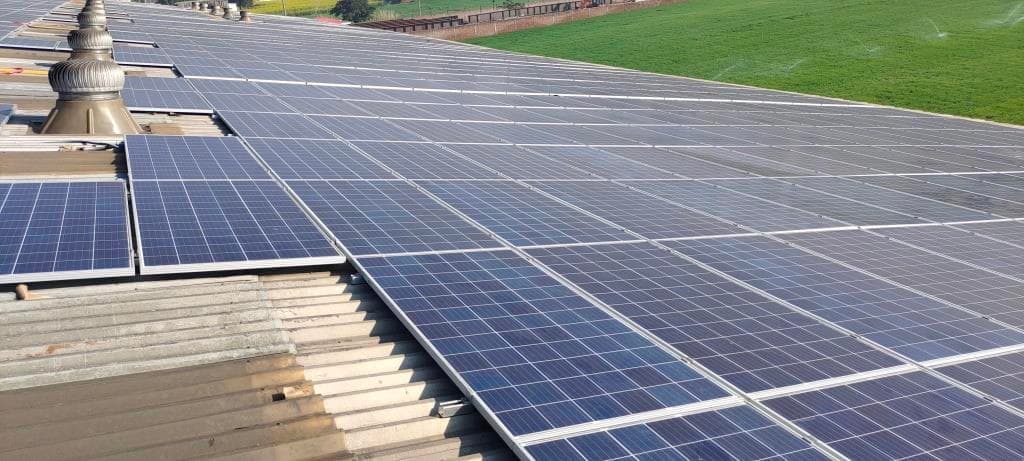 Rows of blue solar modules on a corrugated metal factory roof, two turbine ventilators at the left edge and a green field beyond the roof