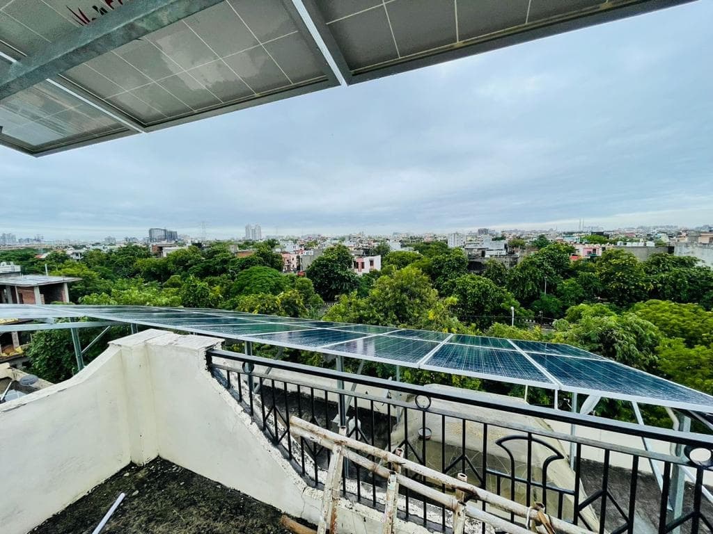 Solar panels on a raised steel structure above a terrace railing, with trees and low-rise houses stretching to the skyline under an overcast sky