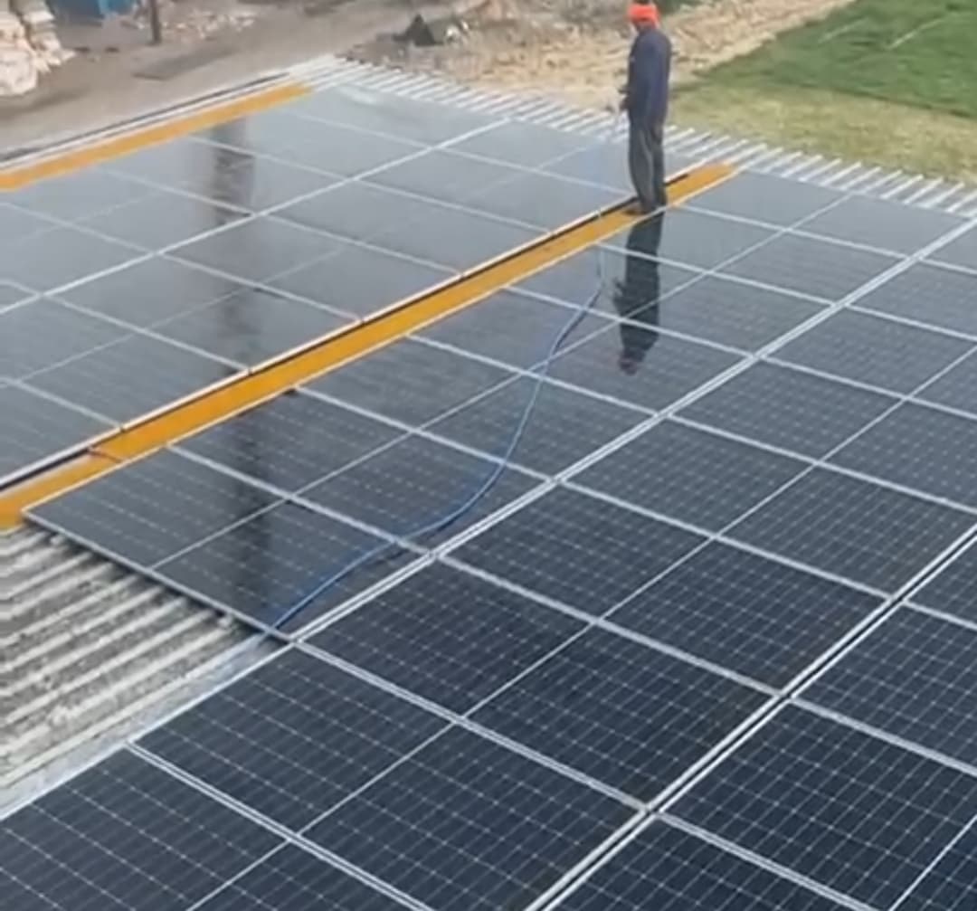 A worker in a red cap stands on a walkway between rows of wet solar panels on a metal-sheet roof, holding a hose, with grass and open ground behind